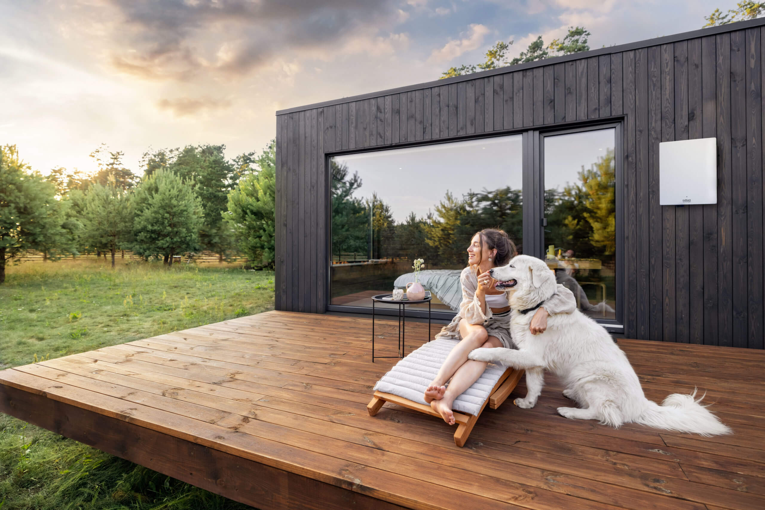 A woman and her dog sit outside her home with a solar inverter on the wall.