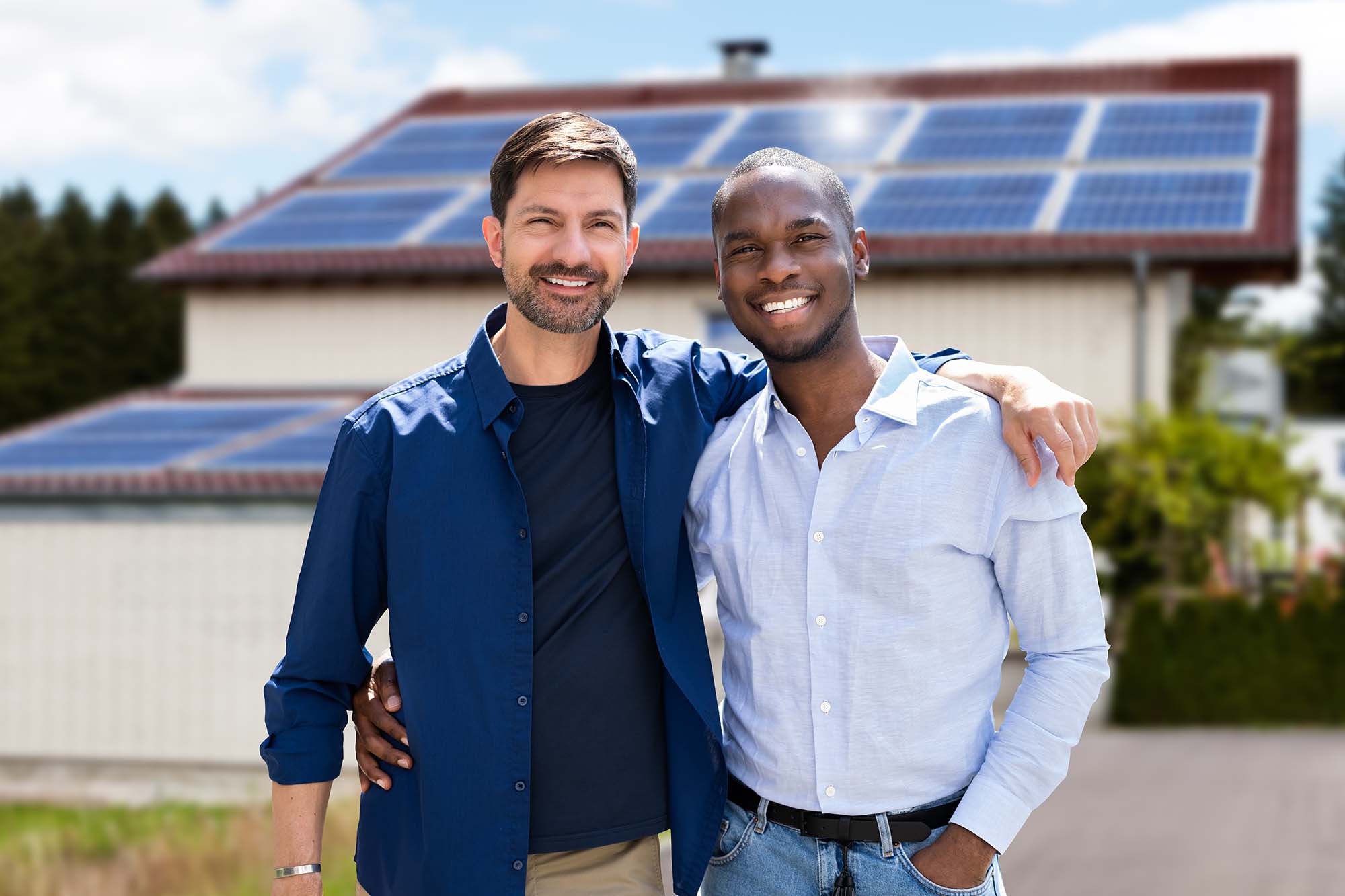 Two men with arms around each other smiling at the camera whilst stood in front of a home with solar panels on the roof.