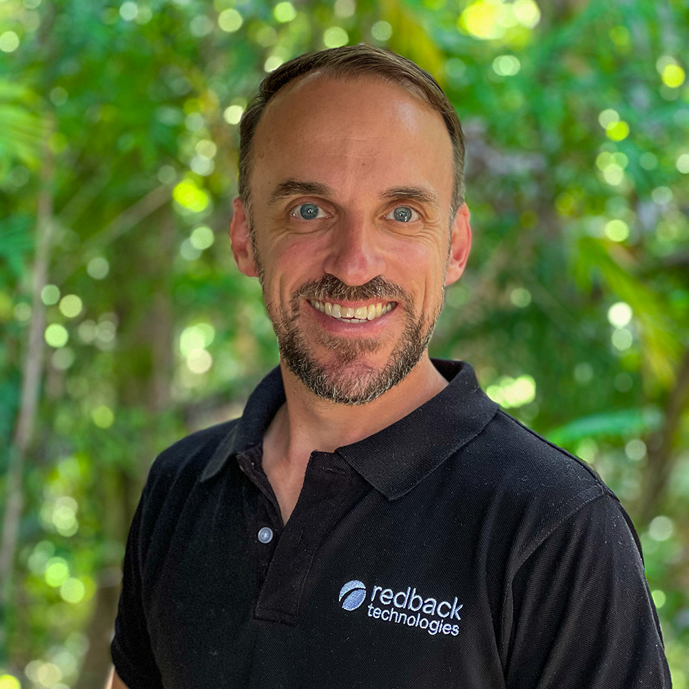 Simon Willis, Redback's Director of Hardware smiling at the camera wearing a black shirt in front of trees