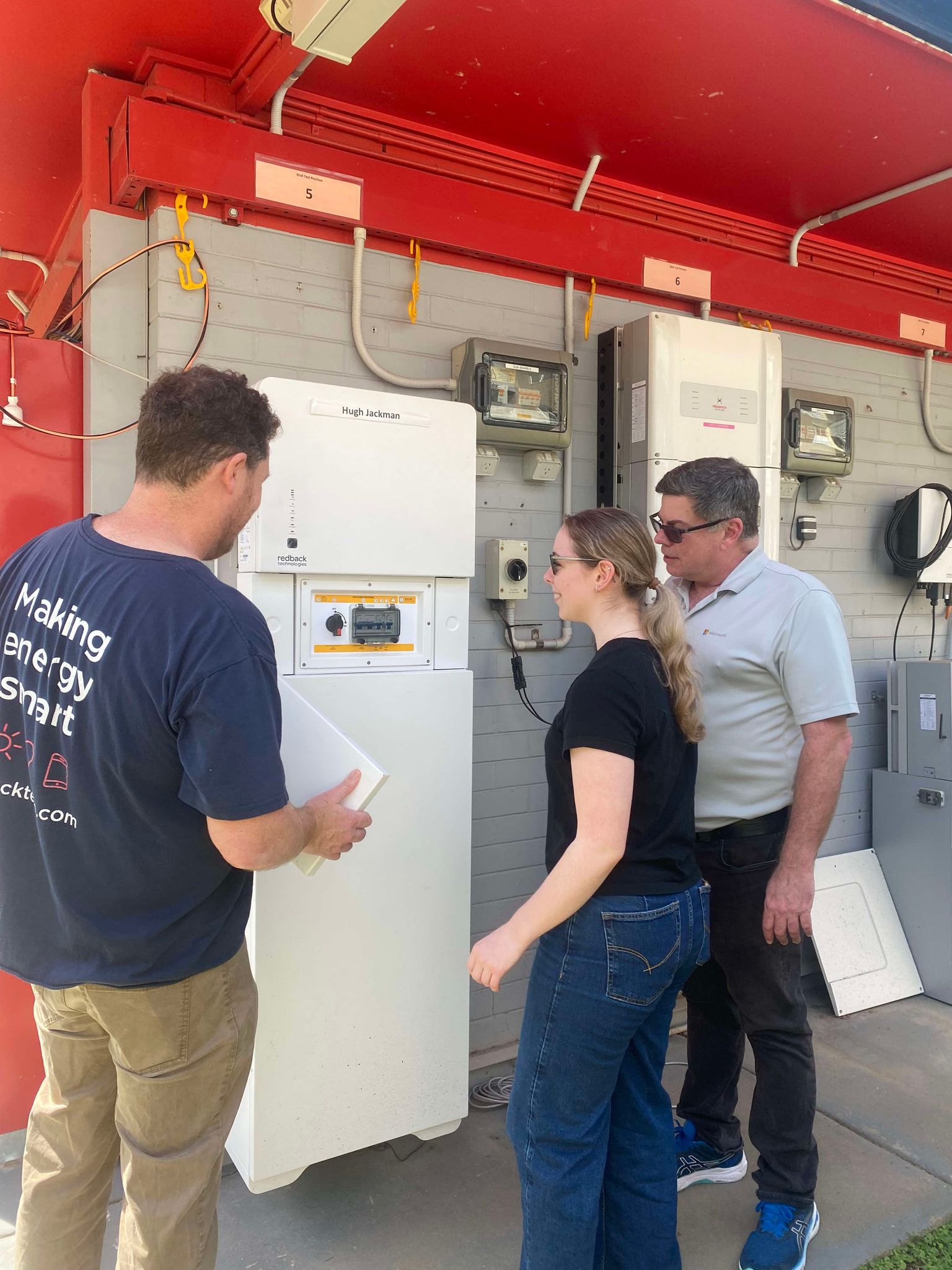 2 men and a woman looking, smiling and pointing at a Redback Hybrid System affixed to a wall